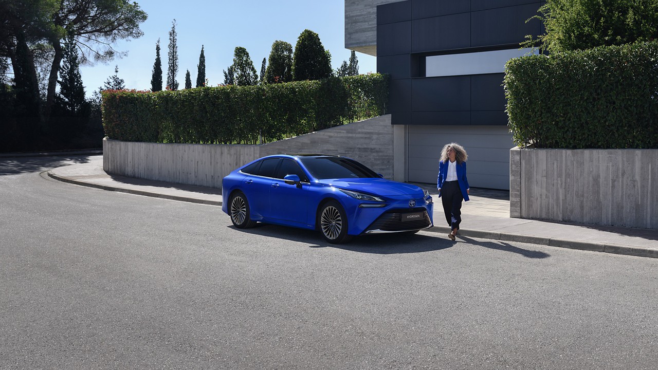 Woman stepping out of a modern house and walking around a bright blue Toyota Mirai, which is parked outside the house on a street