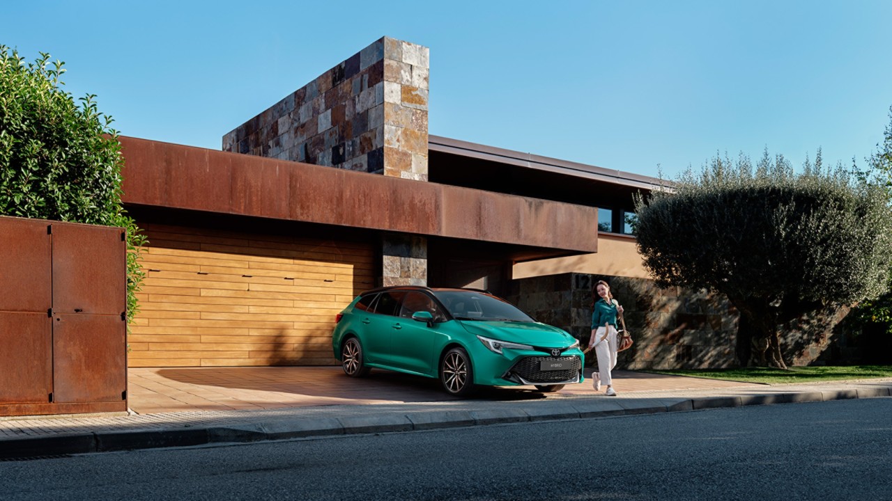 Woman walking out of a modern house, and around a parked green Toyota Corolla