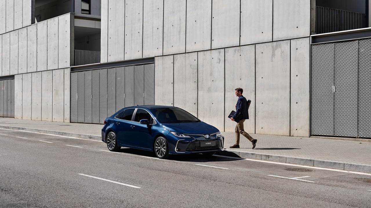 Business man walking towards their navy blue Corolla Sedan, which is parked outside of a modern building