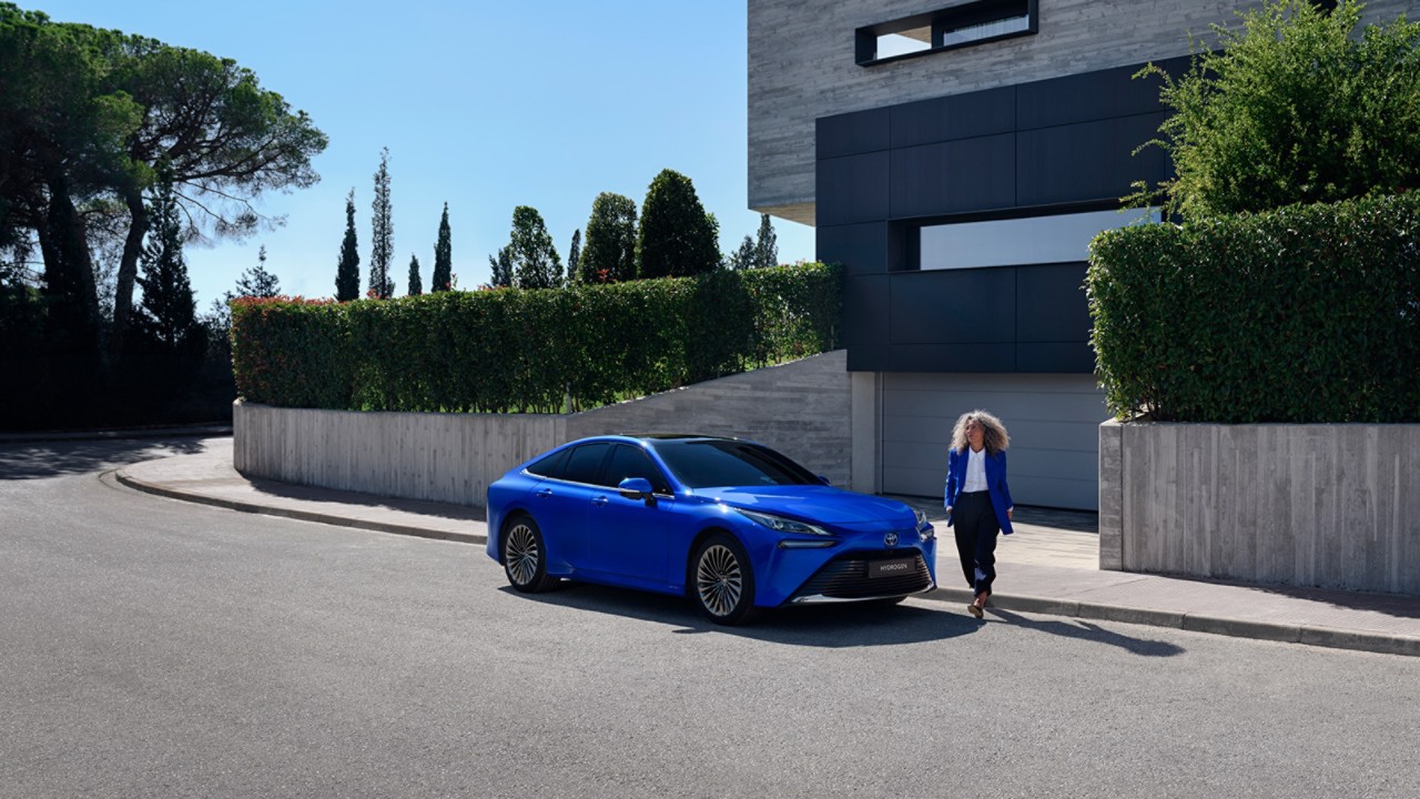 Woman stepping out of a modern house and walking around a bright blue Toyota Mirai.