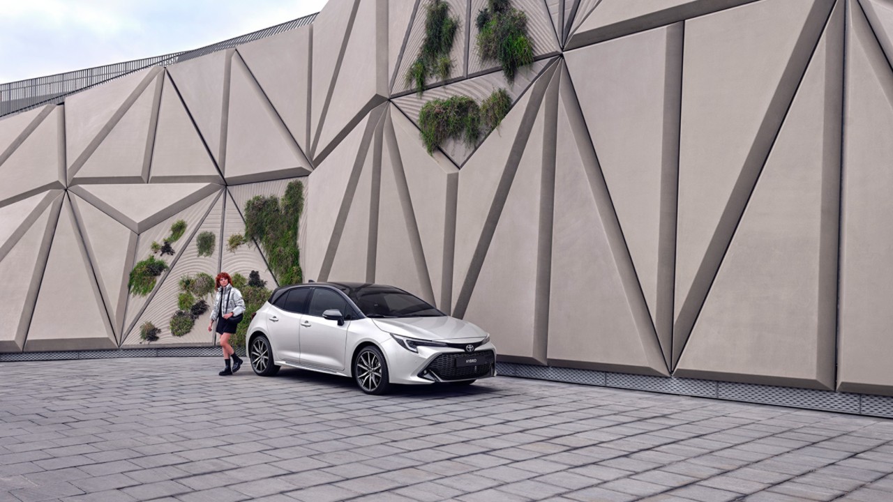 Woman walking outside of a modern abstract building, walking around a silver Corolla Hatchback